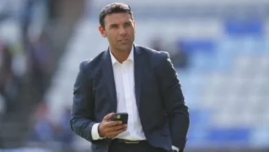 Damián Zamogilny, con traje azul oscuro y camisa blanca, sostiene un teléfono móvil mientras camina en un estadio; su expresión es seria.